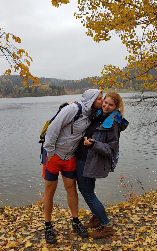 Para całująca się nad jeziorem, otoczona spadającymi liśćmi z jesiennych drzew.