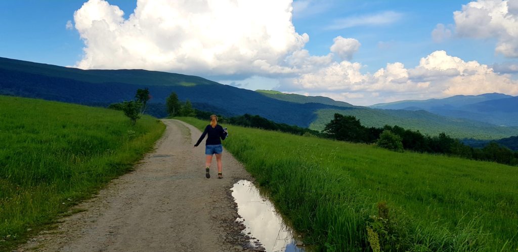 Dziewczyna na bieszczadzkim szlaku otoczona zielona trawą z górami w tle.