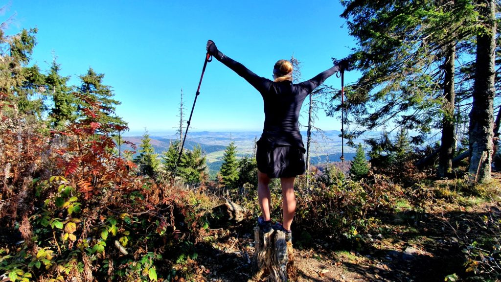 Dziewczyna na pieńku z rozłożonymi rękoma, trzymająca kije trekkingowe i podziwiająca widoki w piękny, słoneczny dzień.