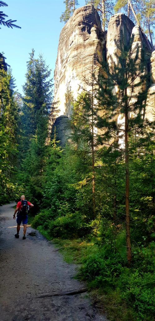 Mężczyzna z plecakiem, idący po leśnej ścieżce obok potężnych skalnych fomacji.
