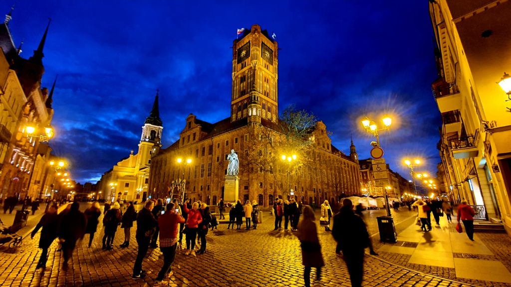 Widok starówki w Toruniu nocą ładnie oświetlony ratusz i ludzie pod pomnikiem Mikołaja Kopernika