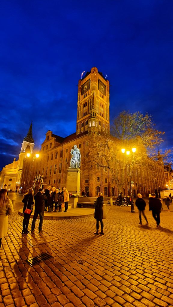 Widok starówki w Toruniu nocą ładnie oświetlony ratusz i ludzie pod pomnikiem Mikołaja Kopernika